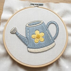 May include: Embroidered watering can design in a wooden embroidery hoop. The watering can is light blue with a yellow flower and white accents. The embroidery is on a light beige fabric.