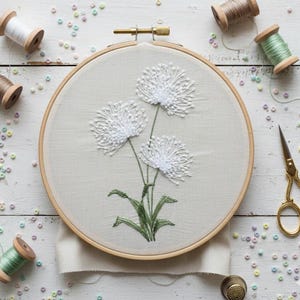 May include: An embroidery hoop with a floral design. The artwork features three white, fluffy flower heads with green stems and leaves, stitched on a beige fabric. Wooden spools of thread, scissors, and beads surround the hoop, suggesting a craft project.