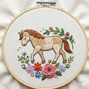 May include: Embroidered horse in a wooden hoop. The horse is light brown with a brown mane and tail, surrounded by a floral wreath of pink, purple, orange, and blue flowers with green leaves. The background is white.