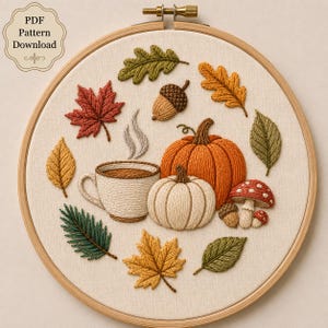 May include: An embroidery hoop containing a fabric panel with stitched autumn motifs including a steaming cup, two pumpkins, an acorn, a mushroom, and various colourful leaves in shades of red, orange, yellow, and green. Text reads PDF Pattern Download.