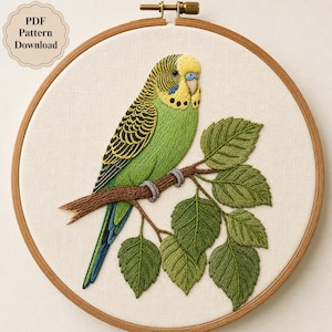 May include: A circular wooden embroidery hoop containing a fabric piece with a hand-stitched budgerigar bird perched on a brown branch with green leaves. The bird features yellow, green, and black plumage. Text in the corner reads PDF Pattern Download.