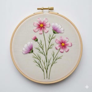 Può includere: Ricamo di fiori di cosmo rosa e bianchi, con centri gialli e steli e foglie verdi, presentato in un telaio di legno. Il motivo floreale è cucito su un tessuto color crema, creando un'opera d'arte artigianale.