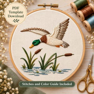 Puede incluir: Un bastidor de bordado con un panel de tela que muestra un pato real bordado en vuelo sobre juncos verdes y agua azul. El texto PDF Template Download y Stitches and Color Guide Included es visible sobre el fondo de tela color crema.