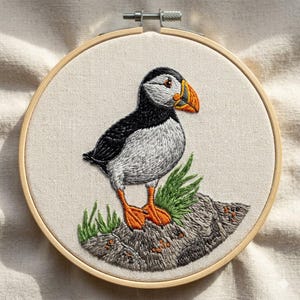 May include: Embroidered puffin bird artwork in a wooden hoop. The bird features black, white, and grey feathers, an orange beak and feet, and is perched on a rock with green grass. The background is a light beige fabric.