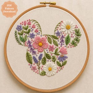 May include: A circular wooden embroidery hoop containing a fabric panel with a floral arrangement stitched in the shape of a mouse head silhouette. The design features pink, white, purple, and green thread depicting various flowers and leaves.