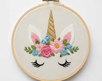 May include: A handcrafted embroidery hoop featuring a unicorn design. The unicorn has a golden horn, white ears with pink interiors, and closed black eyelashes. The design includes pink, blue, and green floral accents, all set against a cream-colored fabric.