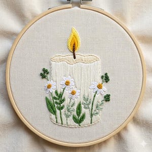 May include: A circular wooden embroidery hoop containing a piece of off-white fabric. The fabric features a hand-stitched design of a cream-coloured candle with a yellow and orange flame, decorated with embroidered white daisies and green leaves.