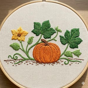 Peut inclure: Un tambour à broder en bois circulaire contenant un panneau en tissu avec un motif brodé à la main représentant une citrouille orange, une fleur jaune et des feuilles vertes sur des vignes, sur un fond en tissu beige clair.