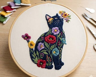 Puede incluir: Diseño de gato bordado sobre tela color crema dentro de un aro de madera. El gato azul oscuro está adornado con bordados florales coloridos, incluyendo amapolas rojas, flores moradas y amarillas. Hilos de bordar y tijeras son visibles en el fondo.