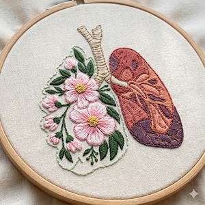 Puede incluir: Un bordado que presenta un diseño de pulmón con detalles florales. El pulmón está representado en tonos rojos y morados, mientras que las flores son rosas con centros amarillos y hojas verdes. La obra está en un bastidor de bordado de madera.