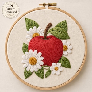 Può includere: Un telaio da ricamo circolare in legno contenente un pannello in tessuto con un disegno ricamato a mano di una mela rosso brillante con un picciolo marrone, circondata da foglie verdi e margherite bianche con centri gialli. Il testo recita PDF Pattern Download.