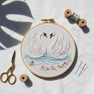 May include: An embroidery hoop with a white fabric featuring two swans facing each other. The swans are white with pink accents, and the water is blue. Gold scissors, thread spools, and needles are also visible.