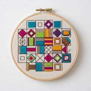 Puede incluir: Un colorido bordado geométrico. El diseño presenta cuadrados, diamantes y otras formas en hilo turquesa, rosa, verde, amarillo y marrón. El bordado está en un aro de madera natural.