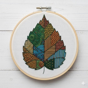 May include: A colorful leaf embroidery in a wooden hoop. The leaf is divided into sections with various patterns and textures, including swirls, stripes, and dots. The color palette includes shades of green, blue, brown, and gold.