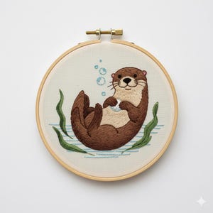 May include: Embroidered otter hoop art. The brown otter is floating on its back, holding a small white object, with bubbles and green seaweed accents. The artwork is set in a wooden embroidery hoop.