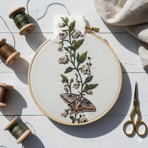 May include: An embroidered hoop art piece featuring a moth and skull design. The artwork is stitched on a cream-colored fabric, with a wooden hoop. The moth has brown and gray wings. Green and brown thread spools and gold scissors are also visible.