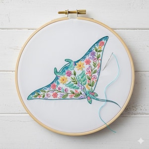 Puede incluir: Un bastidor de bordado con un diseño de manta raya bordado a mano. La raya está llena de coloridos patrones florales en tonos rosa, amarillo y verde. Se ven una aguja y un hilo azul, lo que indica que la pieza está en progreso.
