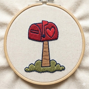 May include: Embroidered artwork in a wooden hoop. The design features a red mailbox with a heart, brown post, and green grass. The embroidery is on a cream-colored fabric.