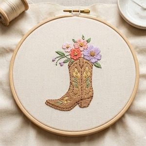 May include: An embroidered artwork featuring a brown cowboy boot filled with colorful flowers in a wooden embroidery hoop. The flowers include pink, orange, and purple blooms with green leaves. The boot has detailed stitching.