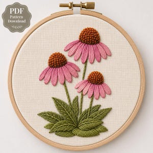Op de afbeelding: Een borduurring met een gebroken witte stof met een handgeborduurd ontwerp van drie zonnehoedbloemen. De bloemen hebben gestructureerde, verhoogde bruine centra en roze bloemblaadjes, met groene bladeren aan de basis. De tekst luidt PDF Pattern Download.