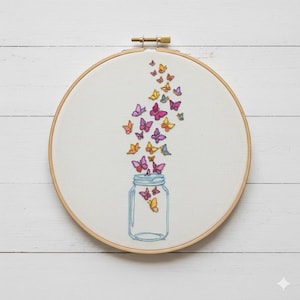 Puede incluir: Arte bordado en un aro de madera. Se representa un tarro azul con mariposas de colores, en tonos rosas, amarillos y naranjas, que emergen de él. El bordado está sobre tela blanca.