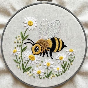 May include: Embroidered artwork featuring a yellow and black bee with white wings, surrounded by white daisies with yellow centers and green leaves. The bee is positioned near the flowers, all set against a light beige fabric background, framed by a gray embroidery hoop.