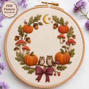 May include: An embroidery hoop containing a fabric piece with a stitched wreath design featuring orange pumpkins, brown acorns, red and white mushrooms, green oak leaves, two small owls, a yellow crescent moon, stars, and a dark red bow.