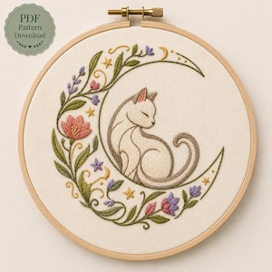 Peut inclure: Un tambour à broder en bois circulaire contenant un panneau en tissu avec un motif brodé à la main représentant un chat crème enroulé en forme de croissant de lune, entouré de vignes vertes, de fleurs roses et violettes et de petites étoiles jaunes.