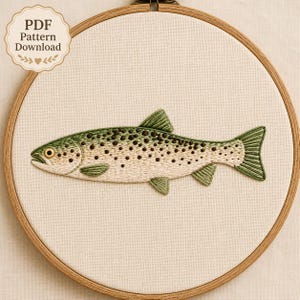May include: A circular wooden embroidery hoop containing a piece of off-white fabric with a hand-stitched trout design. The fish features green and cream-colored thread with dark brown spots. A label in the corner reads PDF Pattern Download.