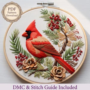 May include: A circular embroidery hoop containing a needlework design of a red cardinal bird perched among green pine needles, brown pinecones, and red berries. The design includes text that reads PDF Pattern Download and DMC & Stitch Guide Included.