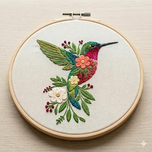 May include: An embroidered hummingbird in a wooden hoop, featuring a vibrant design of green, blue, red, and orange. The bird is surrounded by flowers and leaves, showcasing detailed needlework. The hoop is a standard size, suitable for wall display.