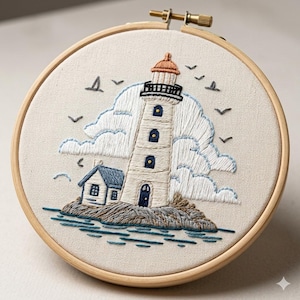 Op de afbeelding: Geborduurd tafereel van een witte vuurtoren met een rood-oranje dak, blauwe ramen en een klein blauw huis. De scène bevat blauwe wolken, vogels en turquoise water, in een houten cirkel.