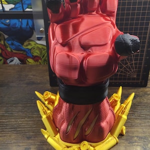 May include: A red and black robotic hand sculpture featuring a metallic finish, positioned upright on a base decorated with yellow bullet-shaped accents. The hand measures approximately 6 inches in height against a black cutting mat background.
