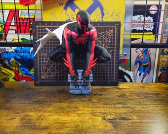 May include: A Spider-Man 2099 figurine in a crouching pose, featuring a black and red suit with a white cape, perched on a gray stone-textured base. The figure is positioned in front of a black cutting mat with visible grid lines and numbers.