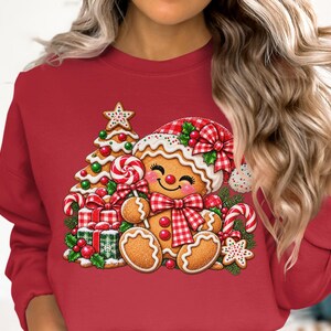 May include: A red crewneck sweatshirt featuring a graphic print of a smiling gingerbread person wearing a red and white chequered hat and bow. The design includes a decorated Christmas tree, wrapped gifts, candy canes, and a star cookie.