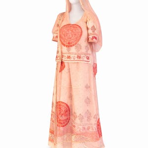 Pode incluir: Um traje indiano tradicional cor de pêssego, incluindo um top, saia e véu, exibido em um manequim. O traje apresenta grandes desenhos circulares vermelhos e padrões intrincados. O tecido parece leve e esvoaçante.