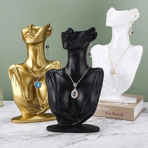 May include: Three jewelry display busts in gold, black, and white. Each bust is sculpted in the shape of a torso and head, and displays a necklace and earrings. The gold bust has a heart-shaped pendant, while the black bust has a teardrop pendant.