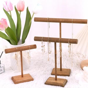 May include: Three wooden jewelry display stands with a T-shaped design. The stands are made of dark brown wood and hold various necklaces. The stands are on a white surface, with a vase of pink and white flowers in the background.