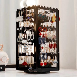 May include: A black, rotating earring display stand filled with various earrings. The earrings are in different styles, including hoops, dangles, and studs, with colors like white, red, and gold. The stand is on a white surface.