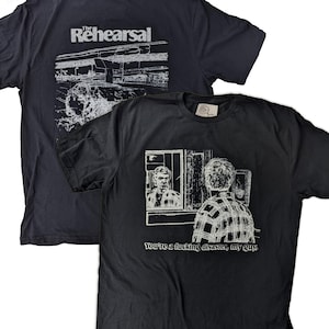 Pode incluir: Duas camisetas pretas. Uma exibe o texto "The Rehearsal" com um gráfico nas costas. A outra camiseta apresenta um gráfico de dois homens e o texto "You're a fucking disaster, my guy."