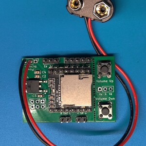 May include: A green circuit board with an SD card slot, buttons labeled "Volume Up" and "Volume Dwn", and various electronic components. A 9-volt battery connector is connected with red and black wires. The background is a solid blue.