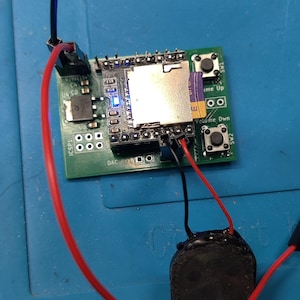 May include: A green circuit board with various electronic components, including an SD card slot, buttons labeled "Volume Up" and "Volume Down", and a small speaker. Red and black wires connect the components.