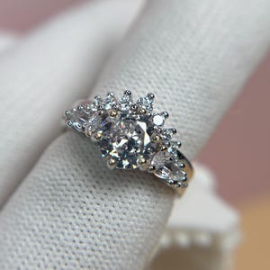 May include: A silver ring with a large, round, clear gemstone at the center, surrounded by smaller, clear gemstones in a floral or starburst pattern. The ring is set on a white surface.