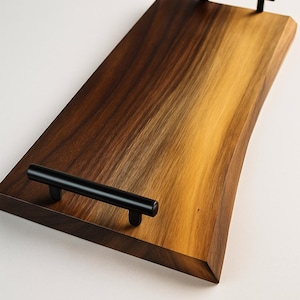 Puede incluir: Una bandeja rectangular de madera con un acabado de vetas naturales. La bandeja tiene un borde marrón oscuro y un centro marrón dorado más claro. Dos asas de metal negro están fijadas a la superficie superior.