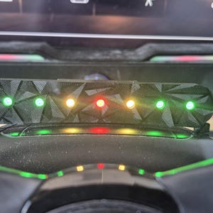 May include: A black, 3D-printed LED indicator strip with a geometric, faceted surface texture. The device features seven circular lights arranged in a horizontal row, currently illuminated in a sequence of green, green, yellow, red, yellow, green, and green.