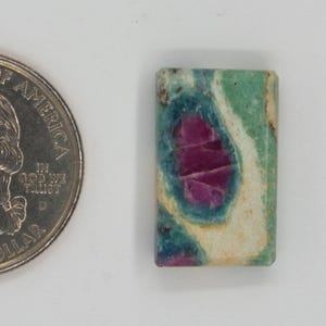 May include: A rectangular gemstone with a vibrant mix of colors, including green, blue, and a prominent magenta center. A U.S. quarter coin is next to the gemstone for size comparison. The gemstone has a unique, natural pattern.