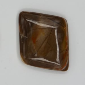 May include: A quarter coin next to a polished, brown gemstone with orange and white inclusions. The gemstone has a rectangular shape with rounded corners. The coin is silver and shows the profile of George Washington.