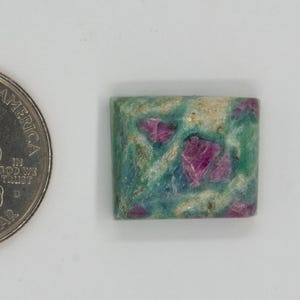 May include: A square gemstone with a mix of green, blue, and cream colors, featuring vibrant red inclusions. A U.S. quarter coin is next to the gemstone for size comparison. The gemstone has rounded corners and a polished surface.