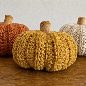 May include: Three crocheted pumpkins in orange, mustard yellow, and cream colors. Each pumpkin features a textured, ribbed stitch pattern and is topped with a small, cylindrical natural cork stem centered at the top of the rounded body.