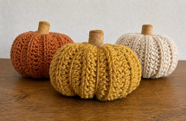 May include: Three crocheted pumpkins in orange, mustard yellow, and cream colors. Each pumpkin features a textured, ribbed stitch pattern and is topped with a small, cylindrical natural cork stem centered at the top of the rounded body.